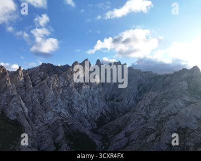 Rugged alpine mountain range with sharp rocky peaks and ridges bathed in sunlight under a vast blue sky dotted with clouds, showcasing dramatic, remot Stock Photo