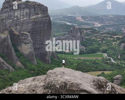 Man standing on a large rock, looking out over the vast and dramatic landscape of meteora, greece, featuring towering rock formations, green valleys, Stock Photo