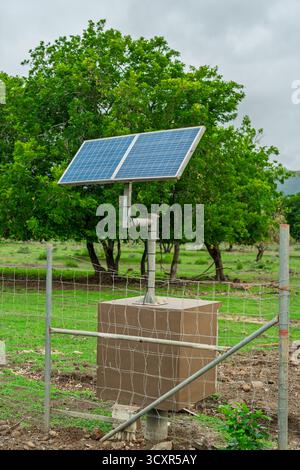 Small blue solar panel array mounted on a post, powering remote monitoring equipment in a fenced wildlife area Stock Photo
