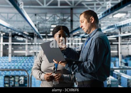 Teamworking data center technicians installing upgraded server racks systems using laptop. Server room coworkers monitoring infrastructure performance using automation tools on notebook Stock Photo