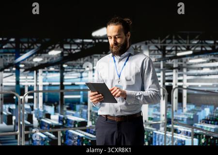 System administrator on data center industrial platform checking code on tablet, managing software updates for server rigs configuration. Male IT professional ensuring data management. Stock Photo