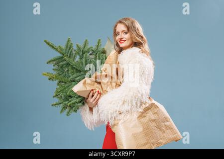Smiling woman in a fluffy coat carries a wrapped evergreen on blue background, perfect for Christmas shopping promos, seasonal greetings, and lifestyl Stock Photo