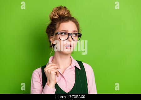 Young woman with glasses wearing a stylish outfit standing against a green background and looking thoughtfully upward Stock Photo
