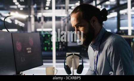 Engineer in data center frustrated by critical error popup on monitor during diagnostics session. IT expert stressed while troubleshooting system fault affecting infrastructure performance, camera A Stock Photo