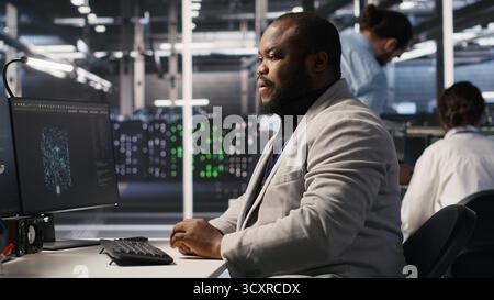 Exhausted server farm employee stretching back after sitting too long at computer desk. Fatigued man in data center office relaxing muscles, stressed by long hardware troubleshooting session, camera A Stock Photo