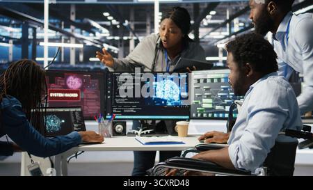 Data center engineer in wheelchair using AI requesting help from coworkers during security breach. Server farm worker with disability aided by colleagues to fight hacker attack stealing data, camera A Stock Photo