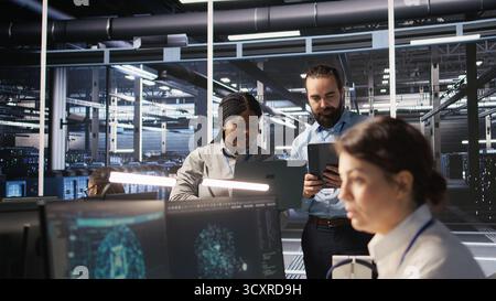 Data scientists working together in server room, reviewing neural network code and LLM visualization dashboards. Team of IT experts coordinating tasks, monitoring AI system performance, camera A Stock Photo