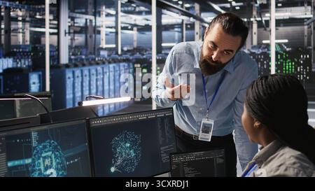 Server room manager providing feedback to employee managing AI training datasets. Team leader talking with technician in data center supervising system operations supporting neural networks, camera A Stock Photo