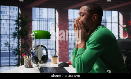 Man feeling exhausted and in pain from lack of sleep at home desk, suffering from insomnia fatigue. Exhausted black guy with negative emotion affected by burnout, feeling stressed. Camera B. Stock Photo