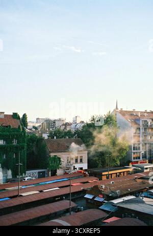 Firefighters extinguish a fire as smoke pours from an old house in the city center, with newer buildings visible behind, captured on analog film. Stock Photo