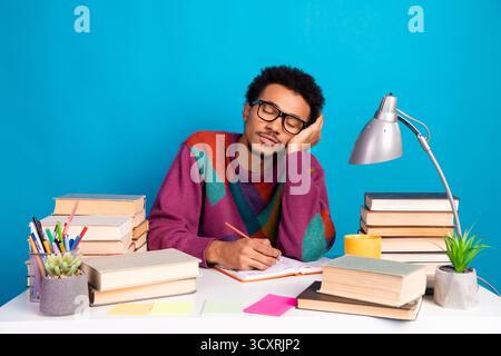 Young man resting his head while studying among stacks of books on a desk against a vibrant blue background Stock Photo