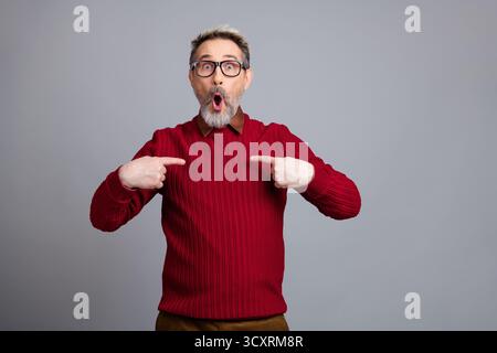 Portrait of a mature man in a red sweater showing an expressive reaction and pointing towards himself on a gray background Stock Photo