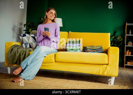 Young woman sits on vibrant yellow sofa in modern living room holding smartphone with folded clothes nearby Stock Photo