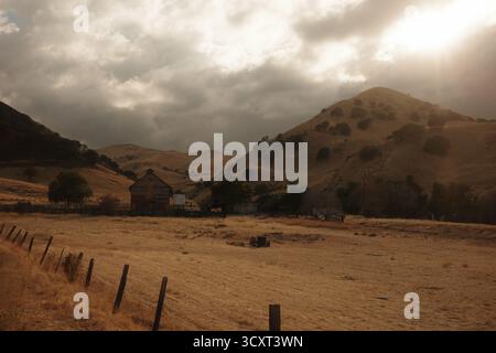 Golden light breaks through storm clouds over an old barn in Black Diamond Mines, casting a warm autumn glow across the rolling hills and dry fields. Stock Photo