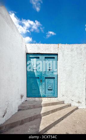 Blue vintage door set against a white stucco wall with steps, under a bright blue sky with scattered clouds Stock Photo