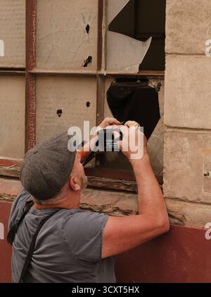 Photographer in flat cap capturing a scene of urban decay through a broken window pane; gritty, melancholic, reflective. Stock Photo