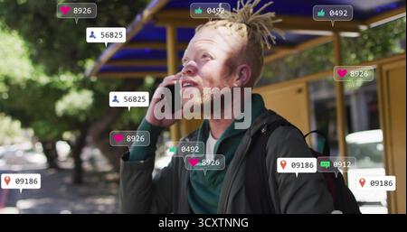 Talking dreadlock man holding smartphone at bus shelter, with floating social media graphics Stock Photo