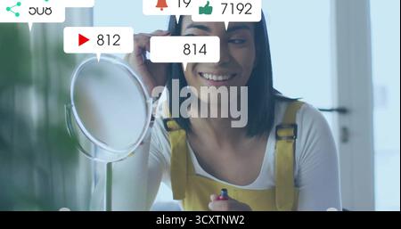 Applying mascara man wearing yellow overalls at home makeup mirror with floating social icons Stock Photo