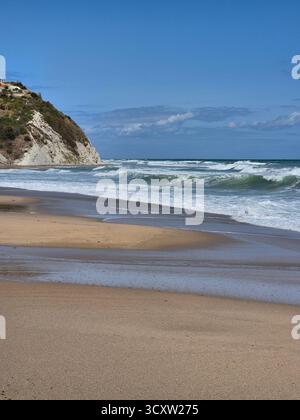 Seascape with a wavy sea rolling onto a sandy shore. In the background on the left, a white or light-colored rock rises above the water. The sky is br Stock Photo