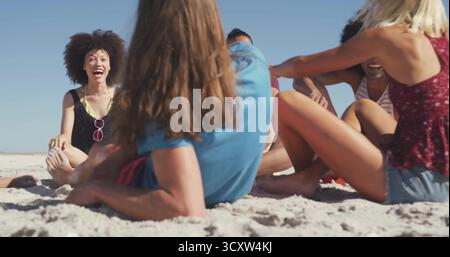 Laughing group of friends sitting in semicircle on white sand at shoreline, with pink sunglasses Stock Photo