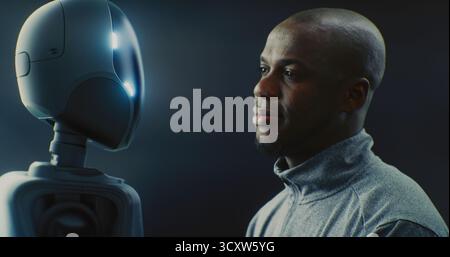 Close Up of African American Man Smiling Warmly While Looking Futuristic AI Robot With Glowing Accents. Positive Human AI Interaction, Friendly Future Of Robotics And Technology Partnership. Portrait. Stock Photo