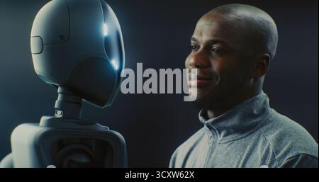 Close Up of African American Man Smiling Warmly While Looking Futuristic AI Robot With Glowing Accents. Positive Human AI Interaction, Friendly Future Of Robotics And Technology Partnership. Portrait. Stock Photo