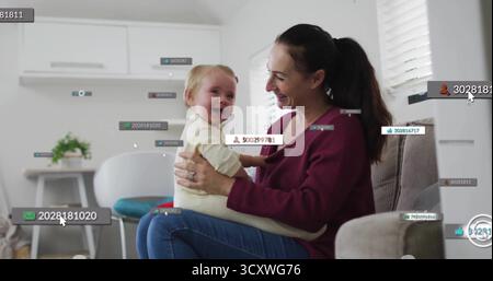 Smiling mother holding baby sharing laughter in living room, with floating digital labels Stock Photo