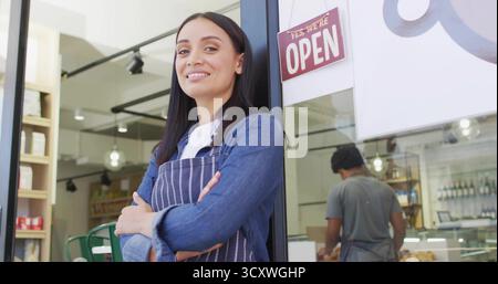Smiling cafe owner wearing denim shirt and striped apron at small cafe entrance, with OPEN sign Stock Photo