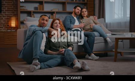 Two diverse married couple four multiracial friends watching television at home party drinking beer alcohol cider drink in living room multiethnic Stock Photo