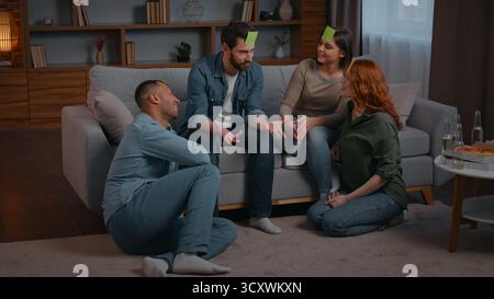 Four diversity ethnic friends playing guessing game stickers on head at home party funny leisure multiracial women men in living room playing charades Stock Photo