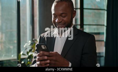 Smiling mature African American businessman using phone chatting mobile app in sunny office business man male executive entrepreneur messaging Stock Photo