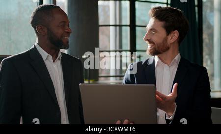 Diverse business team men colleagues businessmen looking laptop happy talking carefree discussing online results partners two multiracial managers in Stock Photo