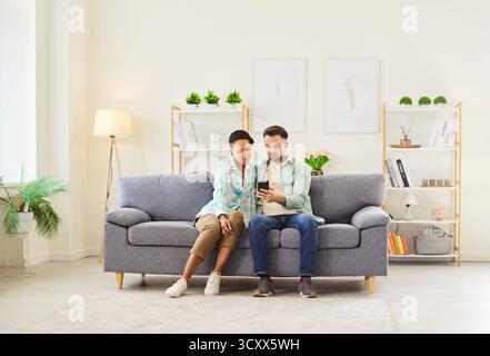 Happy couple sitting on sofa in living room at home and using smartphone together browsing apps. Stock Photo
