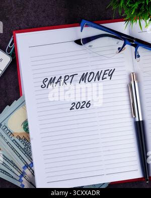 Open notebook displays smart money written boldly on a lined page. Nearby, a pen rests beside stacks of cash and a pair of glasses. A green plant adds Stock Photo