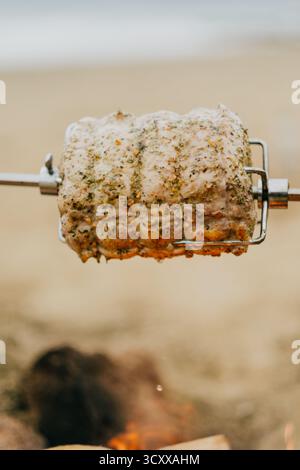 A piece of meat is being cooked on a spit, and it is being cooked on a beach. The meat is being cooked over an open flame, and it is being turned over Stock Photo