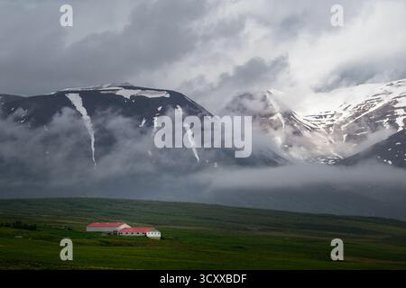 A quaint Icelandic home nestled beneath a snow capped mountain, shrouded in fog and clouds. Stock Photo