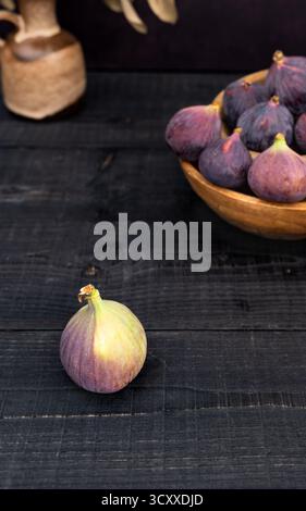 Vertical front view of ripe figs on black wooden table, one fig in focus with bowls of figs behind and vase decor in the background Stock Photo