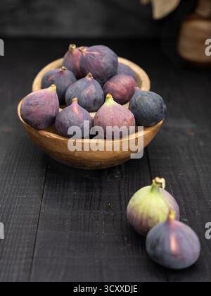 Two ripe figs in front with more in wooden bowls and a vase behind, captured in dark moody style on black wooden background Stock Photo