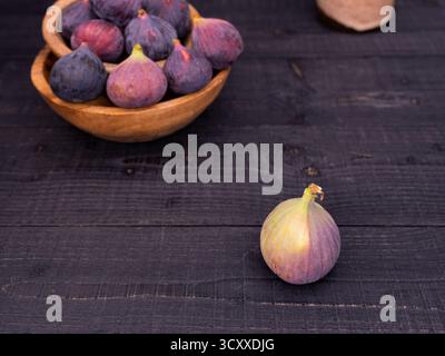 Fresh figs displayed on a black wooden table with one in front and more in wooden bowls, rustic and moody composition with vase in background Stock Photo