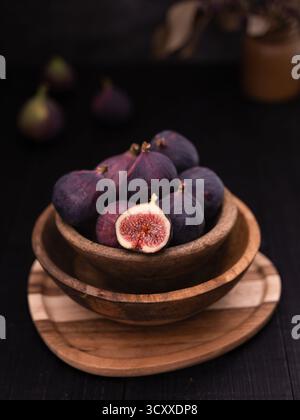 Half sliced fig in wooden bowl on wooden plate with whole figs and vase decor in the background, dark rustic still life Stock Photo
