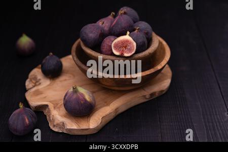 Half sliced fig in stacked bowls on wooden board with whole figs placed diagonally toward background, moody dark still life Stock Photo