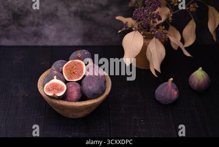 Two sliced figs arranged in a wooden bowl beside whole figs on dark wooden table, moody still life with vase decor in the back Stock Photo