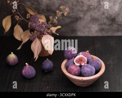Half sliced fig in focus with whole figs nearby in a wooden bowl on dark table, rustic still life with vase decor in background Stock Photo