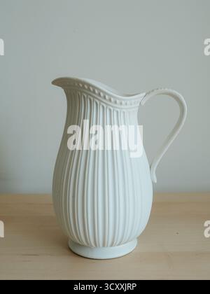 Minimalist photo of a white ceramic pitcher with elegant ribbed texture, placed on a wooden surface against a neutral background Stock Photo