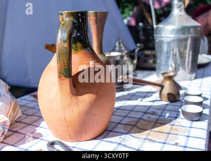A beautiful antique clay jug with a handle standing on a checkered tablecloth near metal utensils and cups at an outdoor historical event Stock Photo