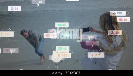 Using smartphone woman in hooded coat framing man at beach with notification icons, copy space Stock Photo