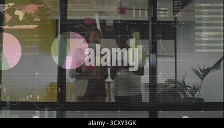 Adjusting businesswomen placing colorful sticky notes on office glass wall, with chart overlays Stock Photo