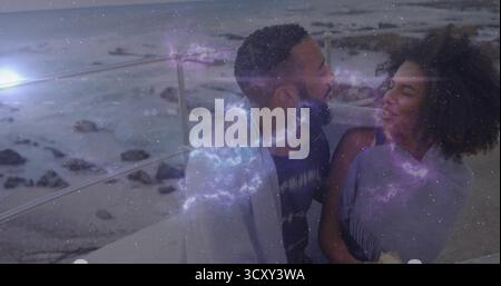 Gazing couple sharing blanket on balcony with metal railing by rocky shore at dusk, cosmic overlay Stock Photo