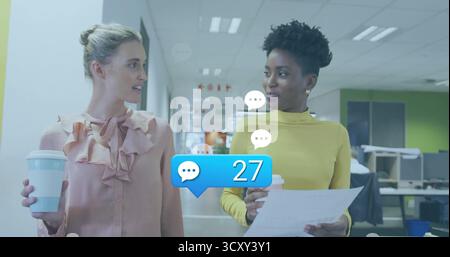 Two colleagues wearing pink blouse, yellow sweater walking in corridor with coffee cups, chat icons Stock Photo