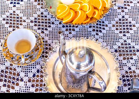 A beautiful still life composition with a silver teapot, golden cup on lace tablecloth and sliced oranges in glass bowl Stock Photo